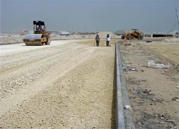 “Construction workers and machinery compacting a gravel base layer along a wide roadbed under development.