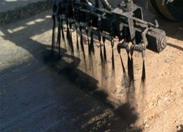 Close-up of spray nozzles applying a dark binder coat over the compacted base layer during road construction.