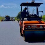 Heavy-duty orange pneumatic road roller parked on a paved surface near equipment storage buildings