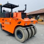 Heavy-duty orange pneumatic road roller parked on a paved surface near equipment storage buildings