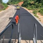 Road construction crew applying fresh black asphalt on a winding road with compactors and workers visible from paver’s perspective