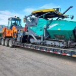 Low-bed trailer transporting a green asphalt paver and an orange road roller to a new project site under blue skies