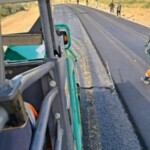Asphalt paver laying a uniform black surface on a rural road while workers manage the edge and finish